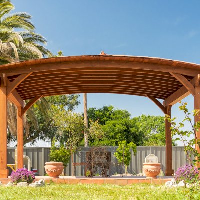 Custom Arched Thick Timber Pavilion (Options: 14' L x 20' Arc W, Mature Redwood, 1 Electrical Wiring Trim, 4-Post Anchor Kit for Concrete, No Ceiling Fan Base, No Privacy Panel, No Curtain Rods, 9 ft H, with 2x6's Roof Boards by Custom Request, Coffee-Stain Premium Sealant). Photo Courtesy of Siino Ventures of Kikrland, Washington.
