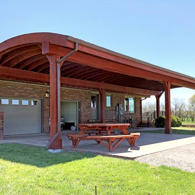 Custom Arched Thick Timber Pavilion (Options: 55' L x 22' Arc W, California Redwood, No Electrical Wiring Trim, 6-Post Anchor Kit for High-Wind, 2 Ceiling Fan Bases, No Privacy Panel, No Curtain Rods, 10ft H, Cherry Stain Premium Sealant) Support timbers covered by custom request. Photo also shows a Round Picnic Table. Photo Courtesy of J. Hartmann of Pittsburg, KS.