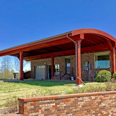 Custom Arched Thick Timber Pavilion (Options: 55' L x 22' Arc W, California Redwood, No Electrical Wiring Trim, 6-Post Anchor Kit for High-Wind, 2 Ceiling Fan Bases, No Privacy Panel, No Curtain Rods, 10ft H, Cherry Stain Premium Sealant) Support timbers covered by custom request. Photo also shows a Round Picnic Table. Photo Courtesy of J. Hartmann of Pittsburg, KS.