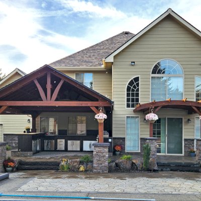 Attached Arched Pergola Kits (Options: 12' L x 12' W, Mature Redwood, Lattice Roof, 2-Post Anchor Kit for High Wind, No Ceiling Fan Base, No Privacy Panels, No Curtain Rods, 10.5' Post Height, Coffee-Stain Premium Sealant). Photo Also Shows a Del Norte Outdoor Kitchen Pavilion. Photo Courtesy of Elements Landscaping of Hayden, ID.