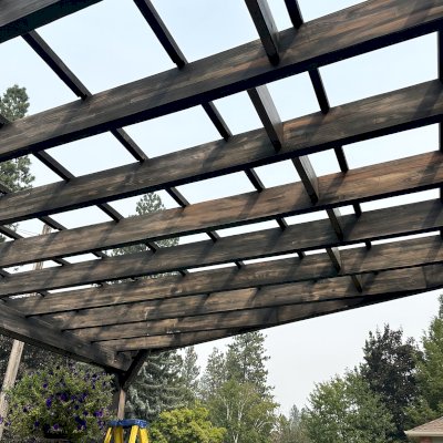 Custom Attached Garden Perglola (Options: Trapezoid Roof Shape [19' Front, 12' Back, 20' Deep], Mosaic Douglas-Fir, Open Roof with Slats at 18", Rafters at 18", with 3 Posts by Custom Request, No Rain Guard, 3-Post Anchor Kit for Concrete, No Privacy Panel, No Ceiling Fan Base, Black Stain Premium Sealant). Photo Taken 1 Year After Installation. Photo Courtesy of T. Perko of Spokane, WA.
