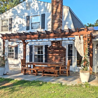Baja Outdoor Redwood Dining Table (Options: 8 ft L, 42 inches W, Side Benches and End Armchairs, Mosaic Redwood, Ruth Chairs, Trestle Benches, 2 Half Length Benches Per Side, No Cushion, Standard Tabletop, Squared Corners, No Umbrella Hole, Transparent Premium Sealant). Photo Also Shows a Traditional Garden Pergola.