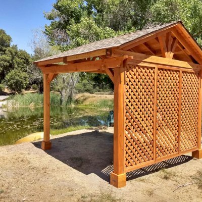 Del Norte Outdoor Kitchen Pavilion (Options: 12' L x 12' W, 1 Full Wall Lattice Privacy Panel, Douglas-fir, 4-Post Anchor Kit for High-Wind, No Ceiling Fan Base, No Electrical Wiring Trim Kit, Straight Internal Truss Supports by Custom Request, No Post Decorative Trims, Transparent Premium Sealant). Photo Courtesy of J. Randall of Warner Springs, CA.