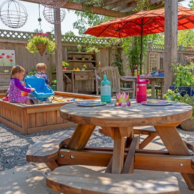 La Playa Sandbox (Options: 96"L x 60" W x 15" H, Mosaic Eco-Wood, Tongue and Grooved Lid, Transparent Premium Sealant). Photo Also Shows a Round Wooden Picnic Table for Toddlers. Photo Courtesy of M. Cooke of Westchester, IL.