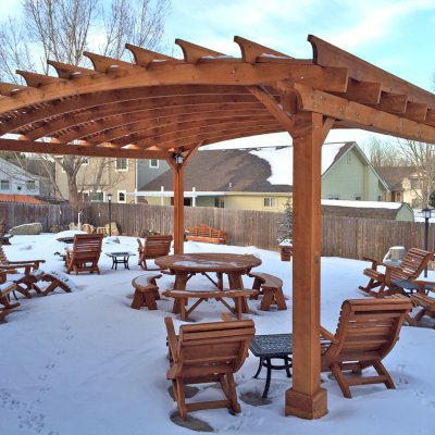 Lutyens Wooden Bench (Options: 8 ft, Mature Redwood, No Cushion, No Engraving, Transparent Premium Sealant). Photo Also Shows an Arched Pergola, a Round Picnic Table Set, 4 Ensenada Easycharis with Ottomans and 4 Ensenada Rocking Chairs in Front of the Bench. Photo Courtesy of Mike Budzinak of Cheyenne, Wyoming.