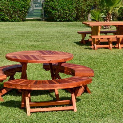 Round Wood Folding Picnic Table (Options: 42", Folding Arc Benches, Redwood [with some Mosaic Redwood boards at the bottom of the seat by custom request], 3 Folding Arc Benches, Standard Tabletop, Checkerboard Design Tabletop, Umbrella Hole & Plug, Transparent Premium Sealant). Photo shows a Trestle Table in Background. 