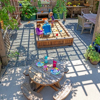 Round Wooden Picnic Table for Toddlers (Mosaic Eco-Wood, No Umbrella Hole, Standard Tabletop, Transparent Premium Sealant). Photo Also Shows a La Playa Sandbox and an Eli's Potting Bench. Photo Courtesy of M. Cooke of Westchester, IL.
