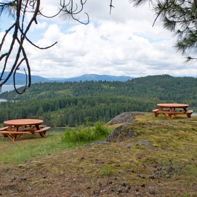 Round Wooden Picnic Tables (Attached Benches) (Options: 6' Diameter, Attached Benches, California Redwood, Standard Tabletop, No Lazy Susan, Umbrella Hole & Plug, Transparent Premium Sealant). These beautiful tables are installed at the “Bull Pine Ledge”, looking out on Lake Pend Orielle from Cloudsledge Conservation Trust, Hope Idaho. Photo Courtesy of Art Green.