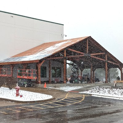 Esther Scissor Truss Pavilion (Options: 33’ L x 63’ W, California Redwood, Standard 3/4" T&G Roof Boards, 16' and 10' H Posts by Custom Request, with 4 Ceiling Fan Bases, High-Wind Post Anchors, 5:12 Roof Pitch, Transparent Premium Sealant). Installation in Process, Photo Shows the Final Roofing Only Partially Installed. Photo Courtesy of the HelloFresh Produce Company of Bolingbrook, IL.