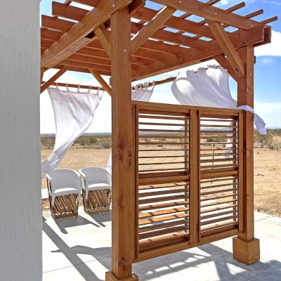 Traditional Wooden Garden Pergola (Options: 12' L x 8' W, Douglas-fir, No Electrical Wiring Trim, Open Roof with Slats at 12", Rafters at 18", with Rain Guard,  4-Post Anchor Kit for High-Wind, No Ceiling Fan Base, 1 Louver Privacy Panel Half Height Without Wall, 2 Curtain Rods, Flush End Cut Detail, 9' Posts, Transparent Premium Sealant). Photo Courtesy of E. Clay of Landers, CA.