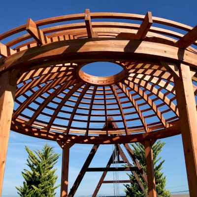 Wooden Dome Pergola (Options: 16' Diameter, with Deck in Coffee Stain, Douglas-fir, Open Roof with Slats at 6", 9' Posts, No Electrical Wiring Trim, 6-Post Anchor Kit for Concrete, Transparent Premium Sealant). Photo Also shows a Round Picnic Table and a Rory's Swing Set in Background.